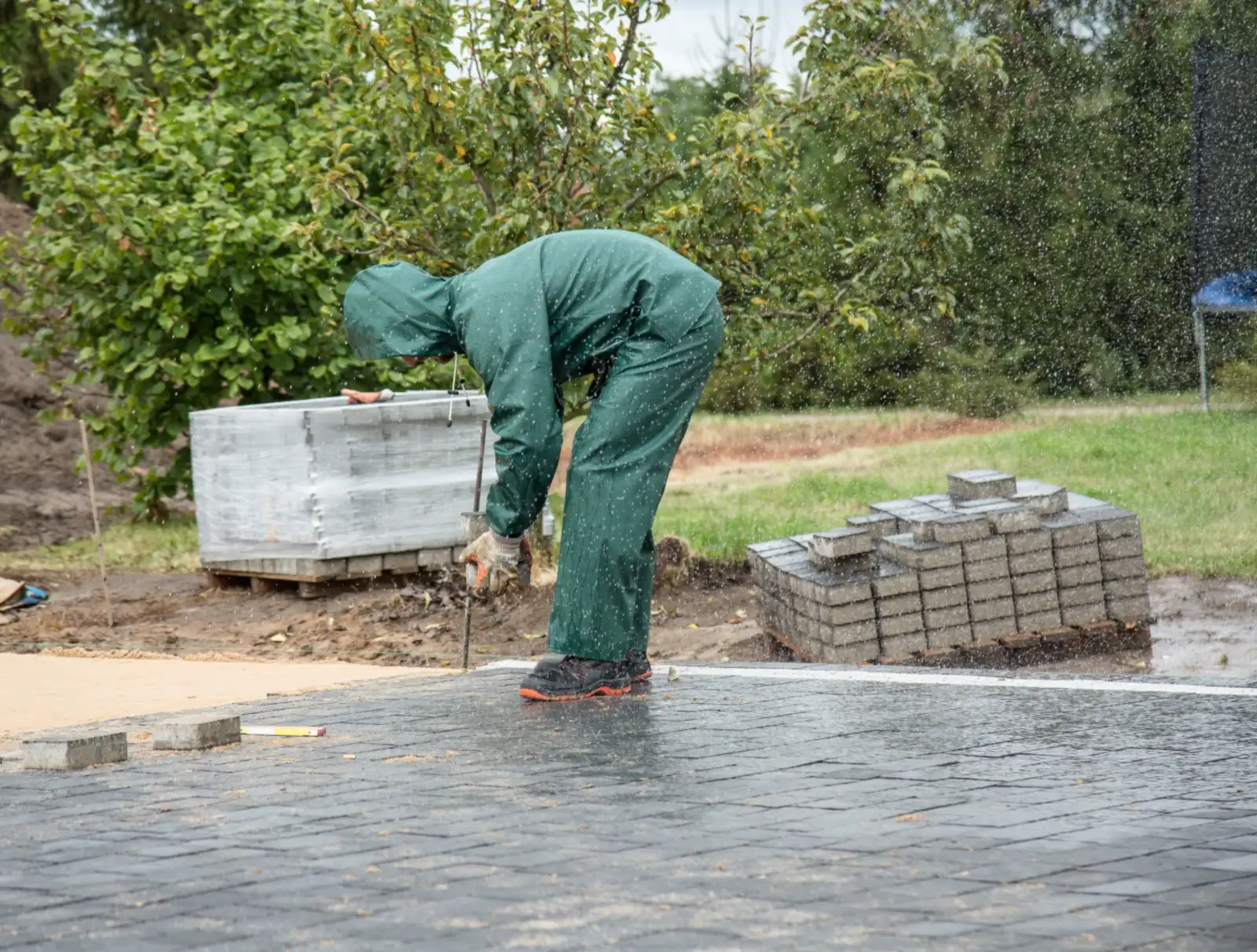 Worker in a wet-weather suit laying pavers in the rain